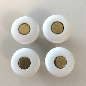 May include: Four white cabinet knobs with a circular design. Each knob has a brass-coloured centre. The knobs are arranged in a square formation, suitable for furniture restoration or new builds.