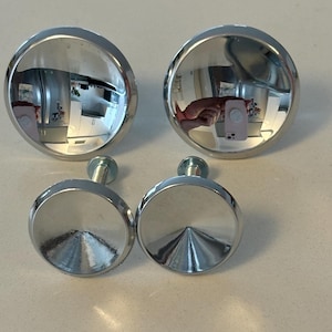 May include: Four shiny, chrome cabinet knobs. Two are rounded with a reflective surface, and two are flat-topped with a conical center. Each knob has a silver screw attached for installation. The knobs are on a white surface.