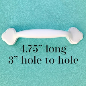 Könnte beinhalten: Weißer Schrankgriff mit Kleeblatt-Enddesign. Der Griff ist 12,07 cm lang, mit einem Lochabstand von 7,62 cm. Der Griff befindet sich vor einem strukturierten, türkisfarbenen Hintergrund.