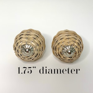 Puede incluir: Dos pomos de armario de ratán tejido con base plateada. Los pomos tienen aproximadamente 4,4 cm de diámetro. Los pomos son de color marrón claro y de forma redonda. Los pomos están sobre un fondo blanco.