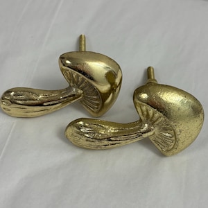 Könnte beinhalten: Zwei goldfarbene Pilz-Schrankknöpfe. Jeder Knopf hat einen Stiel und eine Kappe mit detaillierter Textur. Die Knöpfe sind aus Metall gefertigt und für Möbel oder Dekorationszwecke bestimmt. Die Knöpfe werden auf weißem Hintergrund gezeigt.