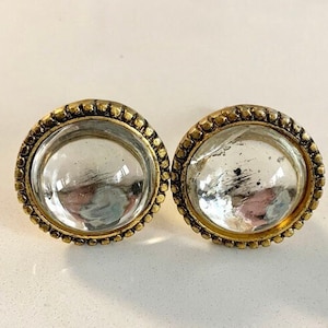 Puede incluir: Un par de pendientes de botón redondos. Cada pendiente presenta un centro transparente y abovedado rodeado por un marco dorado y con cuentas. Los pendientes se muestran sobre una superficie blanca.