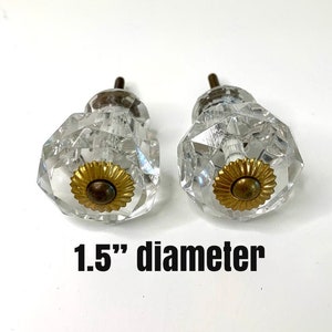 May include: Two clear crystal cabinet knobs with a faceted design and brass-colored bases. The knobs are 1.5 inches in diameter. The knobs are on a white background.