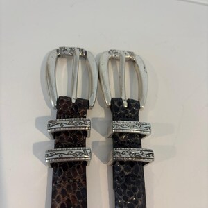 May include: Two leather belts with silver-toned buckles. One belt is brown with a snakeskin pattern, and the other is black with a similar pattern. Both belts have decorative silver accents.
