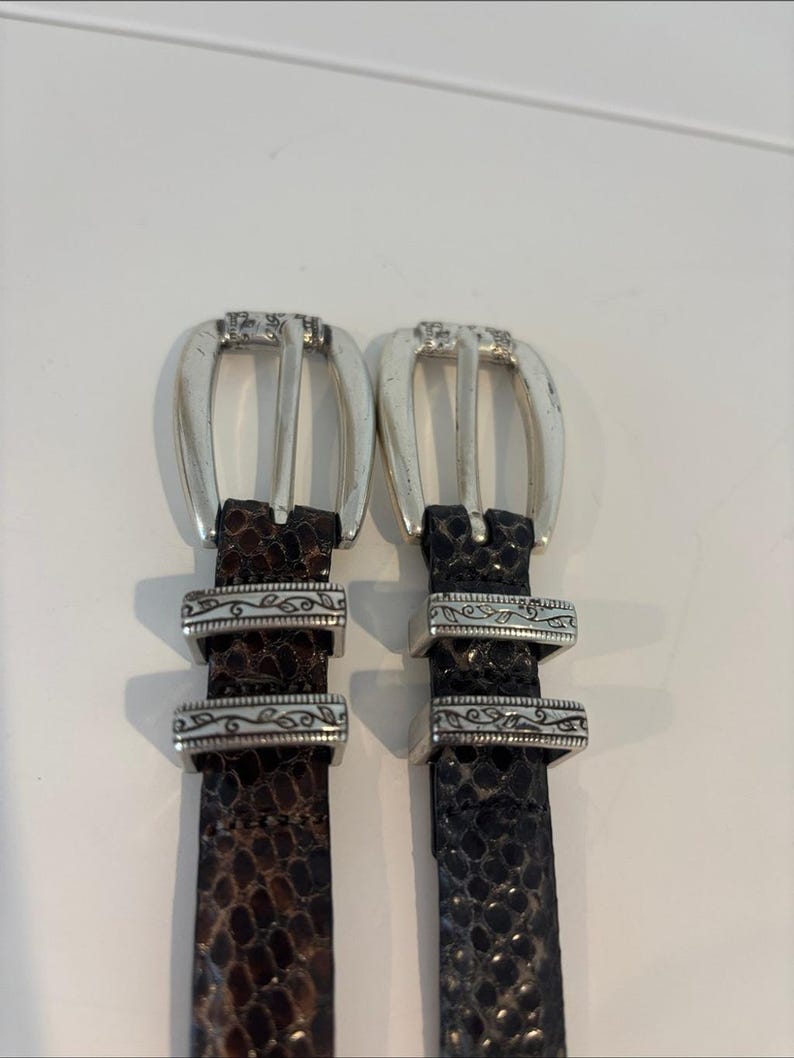 May include: Two leather belts with silver-toned buckles. One belt is brown with a snakeskin pattern, and the other is black with a similar pattern. Both belts have decorative silver accents.