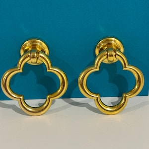 May include: A pair of gold-colored earrings. Each earring features a four-leaf clover design with a circular base. The earrings are set against a teal background.