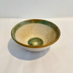 May include: A small, round ceramic bowl featuring a speckled off-white interior and exterior. The rim is accented with a band of muted green and brown glaze, while a circular green patch is centered at the bottom of the bowl's interior basin.