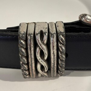 May include: A black leather belt featuring a decorative silver-toned metal loop. The loop consists of five vertical segments, including two twisted rope-style outer pieces, two smooth inner bars, and a central piece with a braided design.