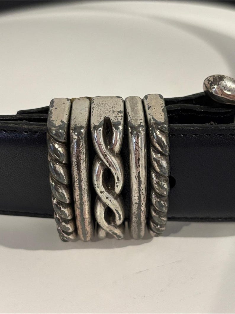 May include: A black leather belt featuring a decorative silver-toned metal loop. The loop consists of five vertical segments, including two twisted rope-style outer pieces, two smooth inner bars, and a central piece with a braided design.