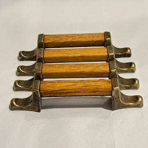 May include: A set of four drawer pulls, each featuring a cylindrical light brown wood center piece held by two aged brass metal brackets with curved feet. The hardware components are arranged in a parallel stack, showcasing the wood grain texture.