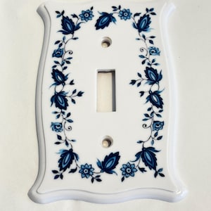 Peut inclure: Une plaque murale d'interrupteur rectangulaire blanche avec un bord festonné décoratif. La plaque présente une découpe rectangulaire centrale pour un interrupteur à bascule et deux trous de vis circulaires. Un motif floral bleu borde le périmètre.
