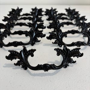 May include: A collection of black metal drawer pulls featuring an ornate, symmetrical design with curved, leaf-like flourishes and intricate detailing. Each handle has a central grip area and two mounting points, arranged in rows on a surface.