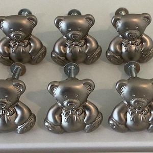 May include: A set of silver teddy bear cabinet knobs. Each knob has a detailed bear design with a bow tie and a screw for fitting. The knobs are arranged in rows, ideal for adding a playful touch to furniture, such as drawers or cabinets.