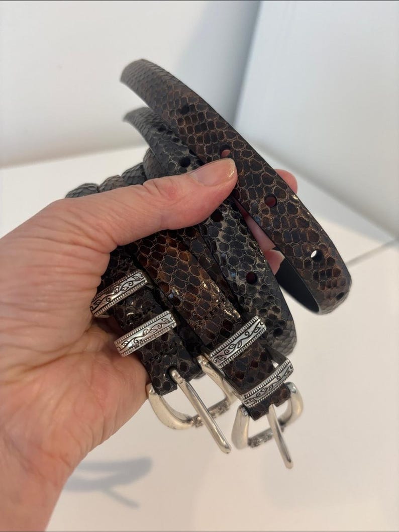 May include: Several brown and black leather belts with silver buckles are held in a hand. The belts have a snakeskin pattern. The buckles have an ornate design.