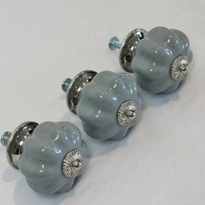 May include: Three light blue ceramic cabinet knobs with a fluted design and silver-toned metal hardware. The knobs have a glossy finish and a decorative silver medallion in the center. Perfect for adding a touch of elegance to furniture.