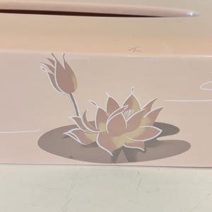 May include: A light pink box with a floral design. The design features a lotus flower and a bud in shades of peach, yellow, and white. The flower is on a brown lily pad. The background is a light pink.