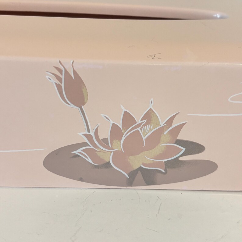 May include: A light pink box with a floral design. The design features a lotus flower and a bud in shades of peach, yellow, and white. The flower is on a brown lily pad. The background is a light pink.