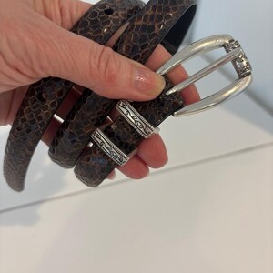May include: A brown snakeskin-patterned belt with a silver-colored buckle. The belt is held up in a hand, showing the texture and design. The buckle has an ornate design.