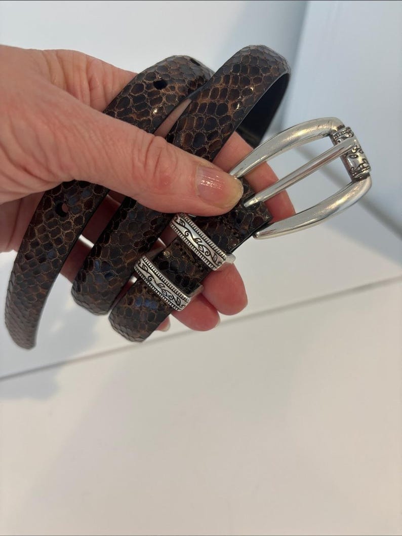 May include: A brown snakeskin-patterned belt with a silver-colored buckle. The belt is held up in a hand, showing the texture and design. The buckle has an ornate design.