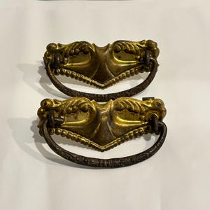 May include: Two matching drawer pulls featuring a gold-toned metal backplate with embossed leaf-like details and a scalloped edge. Each pull includes a dark, textured, semi-circular metal drop handle attached to the central plate with two loops.