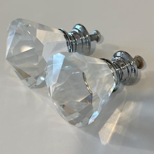 Könnte beinhalten: Zwei klare, facettierte Kristall-Schrankknöpfe mit silberfarbener Metallbeschlägen. Die Knöpfe haben eine rautenförmige Form und reflektieren das Licht. Diese dekorativen Knöpfe eignen sich für Schubladen oder Schränke.