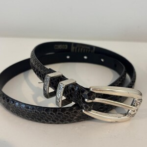 May include: A black belt with a snakeskin pattern and a silver-colored buckle. The belt is coiled on a white surface. The buckle has a decorative design.