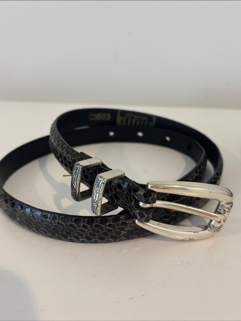 May include: A black belt with a snakeskin pattern and a silver-colored buckle. The belt is coiled on a white surface. The buckle has a decorative design.