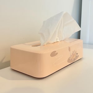 May include: A peach-coloured rectangular tissue box with a floral design of a lotus flower and lily pads. White tissues are pulled from the top opening. The box is on a white surface.