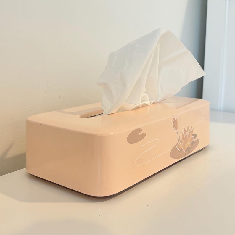 May include: A peach-coloured rectangular tissue box with a floral design of a lotus flower and lily pads. White tissues are pulled from the top opening. The box is on a white surface.