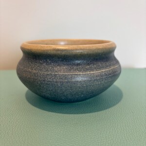 May include: A small, handcrafted ceramic bowl with a rounded shape. The bowl has a speckled, dark blue exterior and a light tan rim. The interior is a lighter tan colour. The bowl sits on a light green surface.