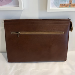 May include: A rectangular brown document case with a textured finish. The case features a horizontal zippered pocket across the center with a tan fabric lining and a metallic zipper pull. A larger zipper closure runs along the top edge of the case.