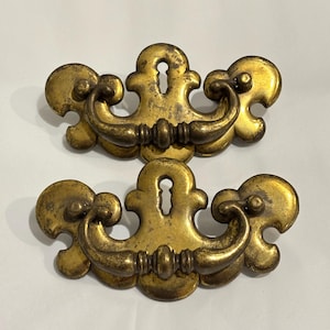 Könnte beinhalten: Zwei passende Schubladengriffe aus Messing mit verwittertem Gold-Finish. Jedes Stück verfügt über ein zentrales Schlüsselloch-Schild mit dekorativem Wellenrand und einen geschwungenen, beweglichen Bügelgriff, der an zwei runden Befestigungspunkten sitzt.