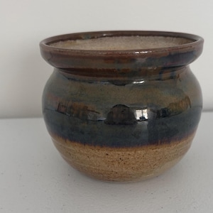 Puede incluir: Una olla de cerámica hecha a mano con un cuerpo redondeado y un borde ligeramente ensanchado. La olla presenta un degradado de color, que va desde una base de color beige claro hasta una sección media azul-verdosa oscura y un borde marrón oscuro. El interior es de color beige claro.