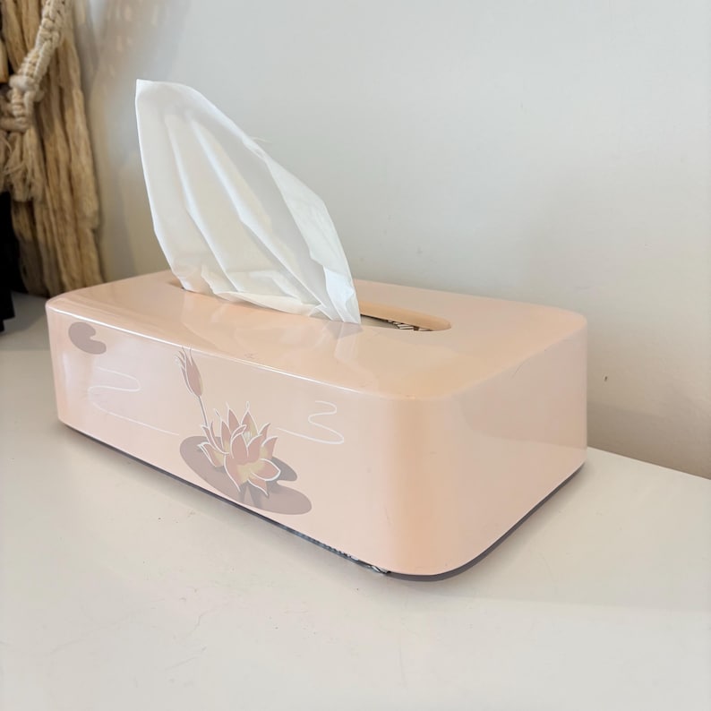 May include: A peach-coloured rectangular tissue box with a floral design of a lotus flower and lily pads. White tissues protrude from the top opening. The box is on a white surface.