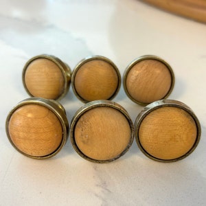 May include: Six wooden cabinet knobs with brass-colored metal rims. The knobs have a light brown, circular wooden center. The knobs are arranged in two rows of three, with a slight angle. The background is a light-colored surface.