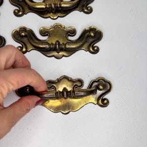 May include: Antique brass drawer pulls with ornate detailing. The pulls have a curved handle and decorative backplate. The hardware is a warm, aged brass color, perfect for vintage furniture restoration projects.