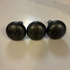Peut inclure: Trois boutons de meuble gris foncé avec une finition mouchetée. Chaque bouton est rond avec une petite vis métallique. Les boutons sont disposés en rangée sur une surface claire.