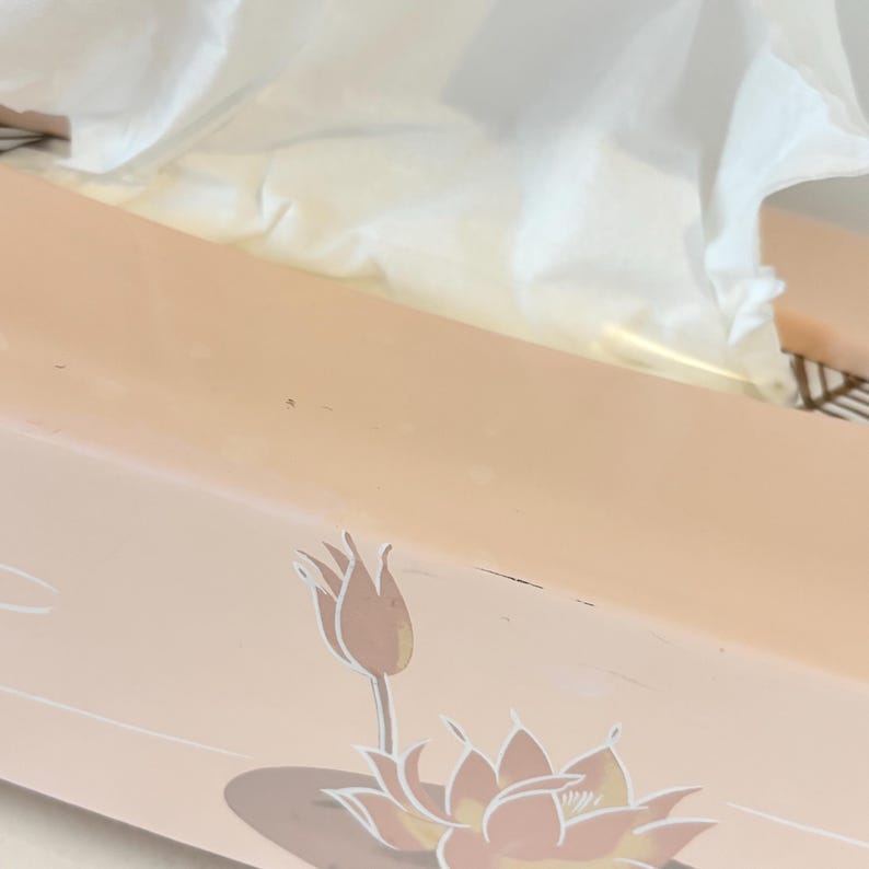May include: A peach-coloured rectangular tissue box with a lotus flower and bud design in peach and white. White tissues are visible at the top. The box is designed to hold tissues.