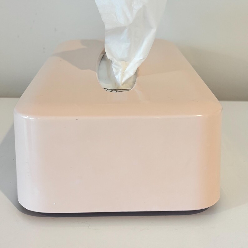 May include: A rectangular, pale pink tissue box with a glossy finish. White tissues protrude from a circular opening on the top. The box is on a white surface.