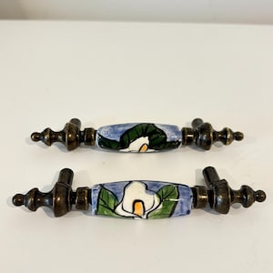 May include: Two decorative cabinet pulls with a floral design. The pulls have a bronze finish with a ceramic center featuring a white calla lily with green leaves against a blue background. The pulls are approximately 15 cm long.