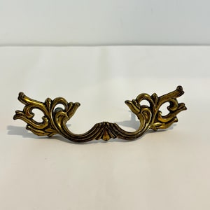 May include: A brass-toned metal drawer pull featuring an ornate, symmetrical design with curved, scrolling flourishes on both sides. The hardware has a weathered, antique finish with darker recessed areas and a central mounting point.