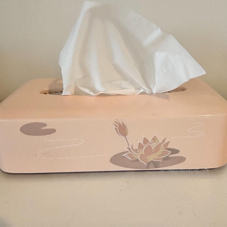 May include: A peach-coloured rectangular tissue box featuring a lotus flower and lily pad design. White tissues are protruding from the top. The box is placed on a white surface.