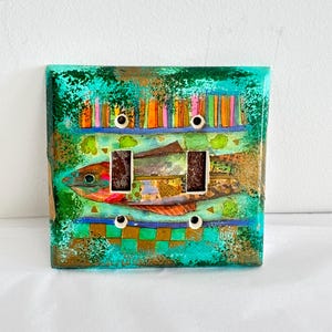 May include: A rectangular double light switch plate featuring a vibrant, multi-coloured collage design. The artwork depicts a stylised fish in shades of red, green, and gold, set against a textured teal background with vertical stripes and a checkered pattern.