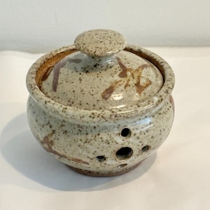 Può includere: Una ciotola in ceramica rotonda per filati con coperchio abbinato, caratterizzata da una smaltatura beige maculata con accenti marroni. La ciotola presenta un'apertura circolare sul lato per il filo e un piccolo pomello sul coperchio.