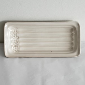 May include: An off-white ceramic serving tray, rectangular in shape. It has a raised rim and a textured surface with horizontal lines. One end features a cluster of small, raised decorative elements.
