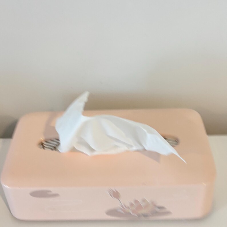 May include: A rectangular, pale pink tissue box with a floral design. White tissues are pulled from the top. The box is on a white surface. The box is likely made of plastic.