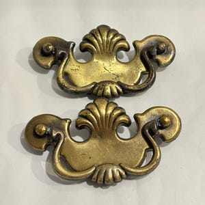 Puede incluir: Dos tiradores de cajón de latón a juego con un motivo central de concha y asas curvas oscilantes. El metal tiene una pátina envejecida y desgastada con arañazos visibles y zonas oscurecidas, mostrando un diseño clásico y ornamentado.
