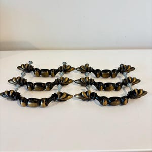May include: A set of six antique-style drawer pulls. Each pull has a curved, dark bronze body with decorative leaf-like accents. They are mounted with silver screws.
