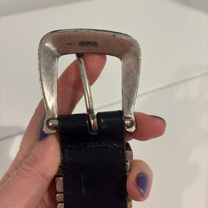 May include: A black leather belt featuring a silver-toned metal buckle with a weathered finish. The buckle displays the embossed text GUESS at the top center. The belt strap is made of smooth black material and connects to the metal hardware.