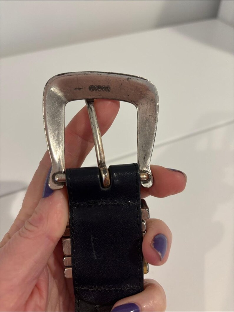 May include: A black leather belt featuring a silver-toned metal buckle with a weathered finish. The buckle displays the embossed text GUESS at the top center. The belt strap is made of smooth black material and connects to the metal hardware.
